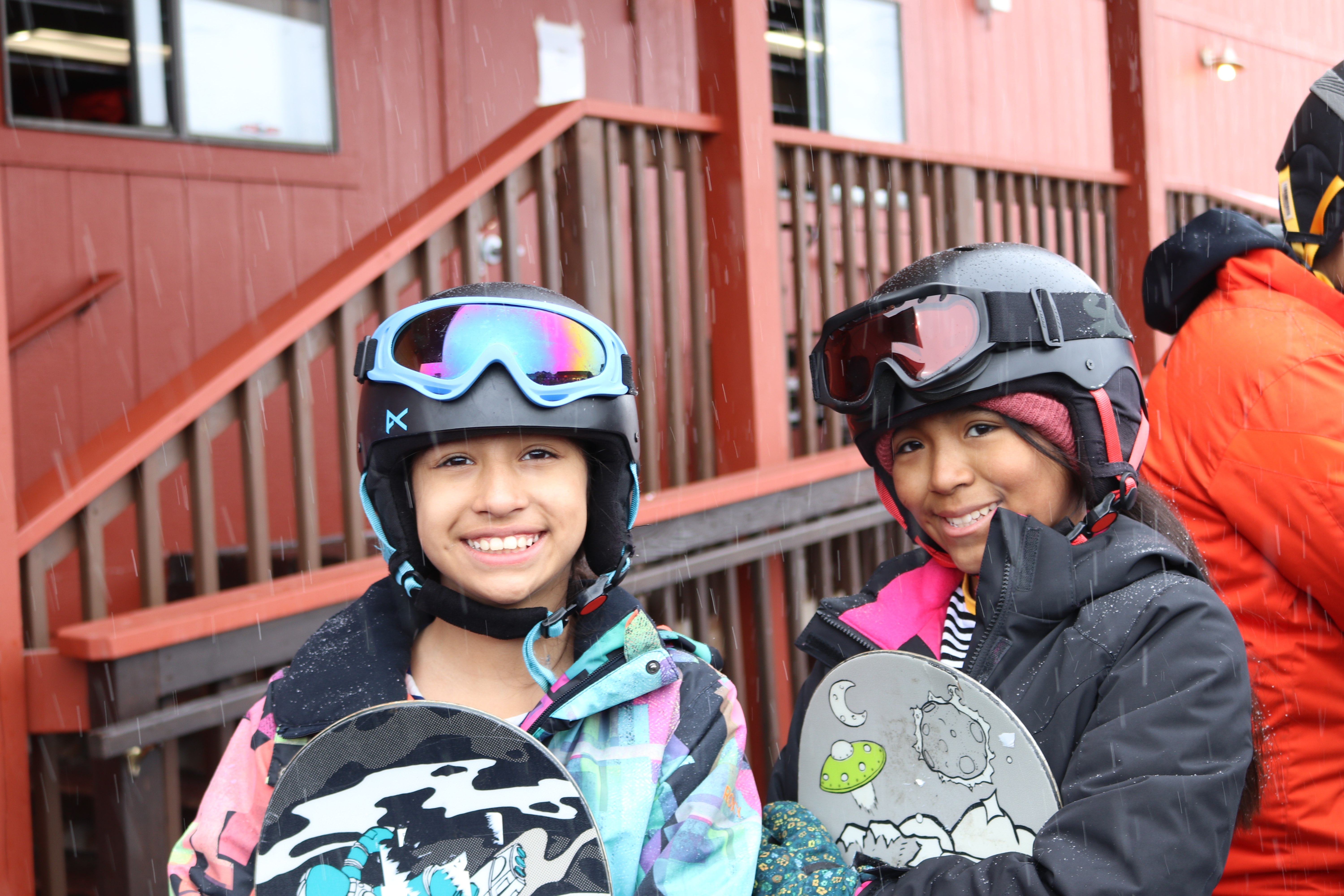 two girls hold snowboards and wear helmets and goggles on their heads